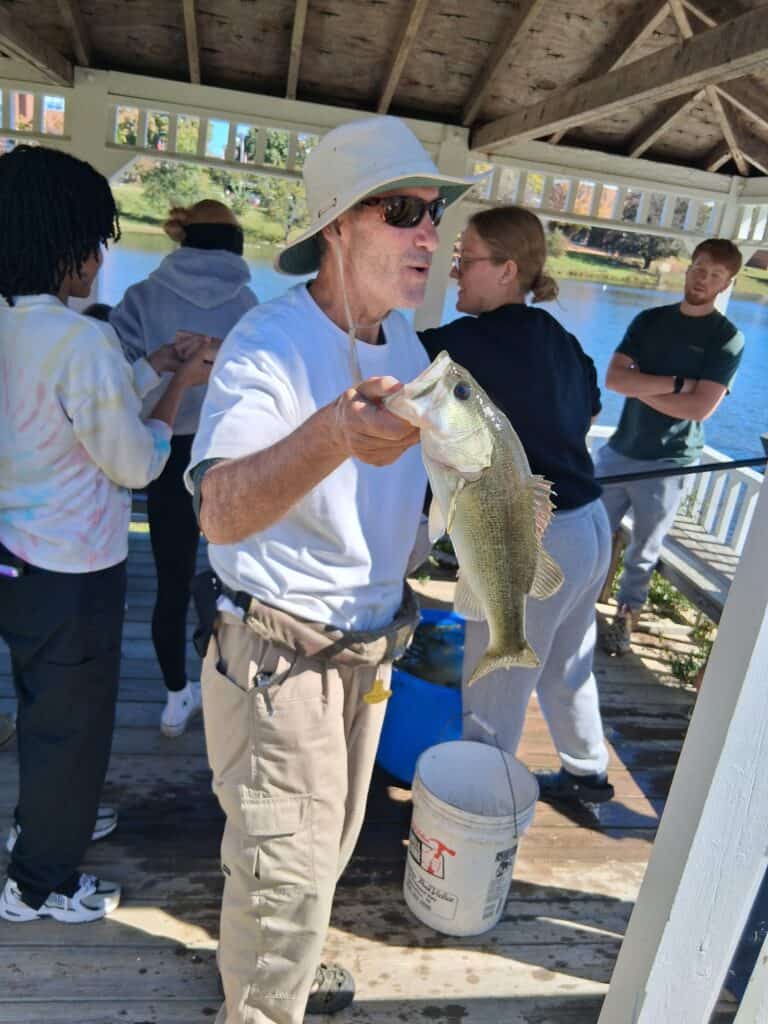 A man in a sun hat holds a largemouth bass on a wooden pier, surrounded by several people in a gazebo overlooking a lake on a sunny autumn day.