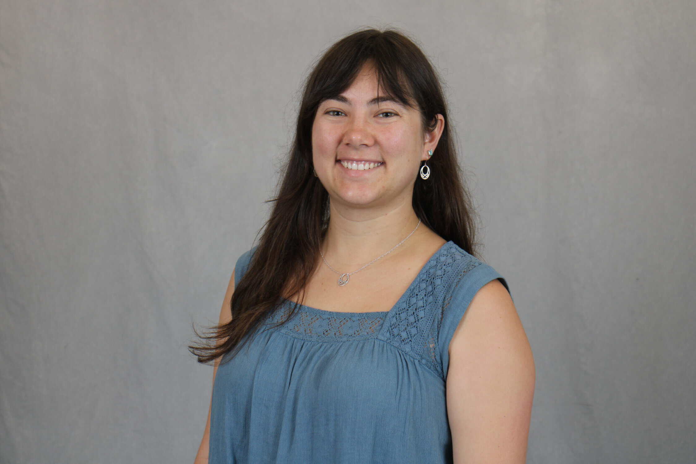 Headshot of woman with long dark hair, dark turquoise sleeveless blouse, grey background
