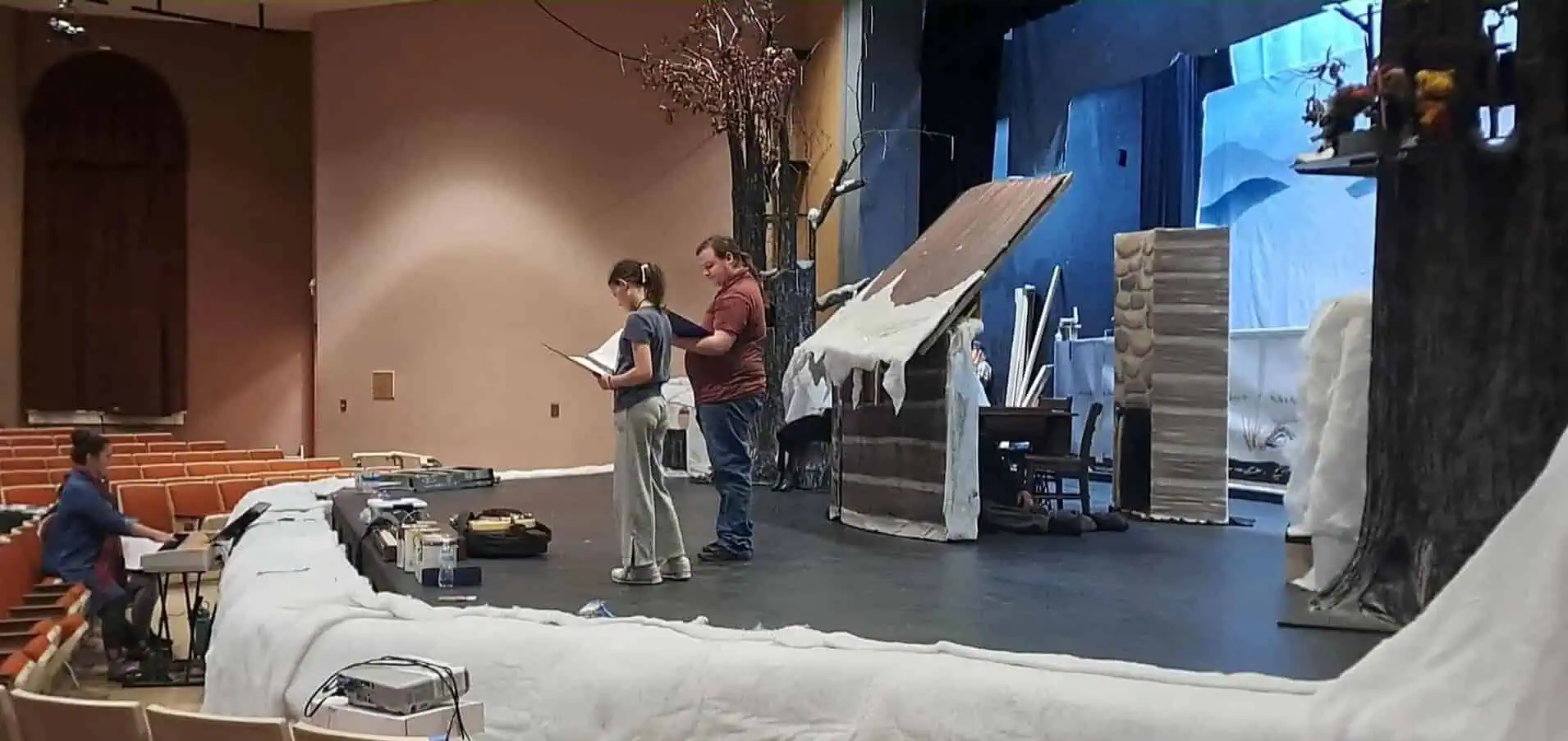 A young man and a girl on stage in a winter setting rehearse the "Emmet Otter" play with Director Emily Blankenship-Tucker playing keyboard.