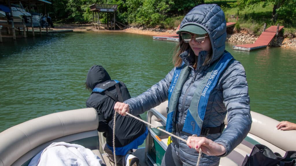 A woman in a gray jacket and green cap is holding a rope on a boat, while another person is leaning over the side near the water.