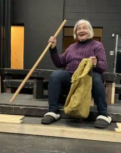 Actor Becky Prophet in purple sweater and slacks with cloth sack and walking stick, rehearsing in black-box theatre sitting on a low platform