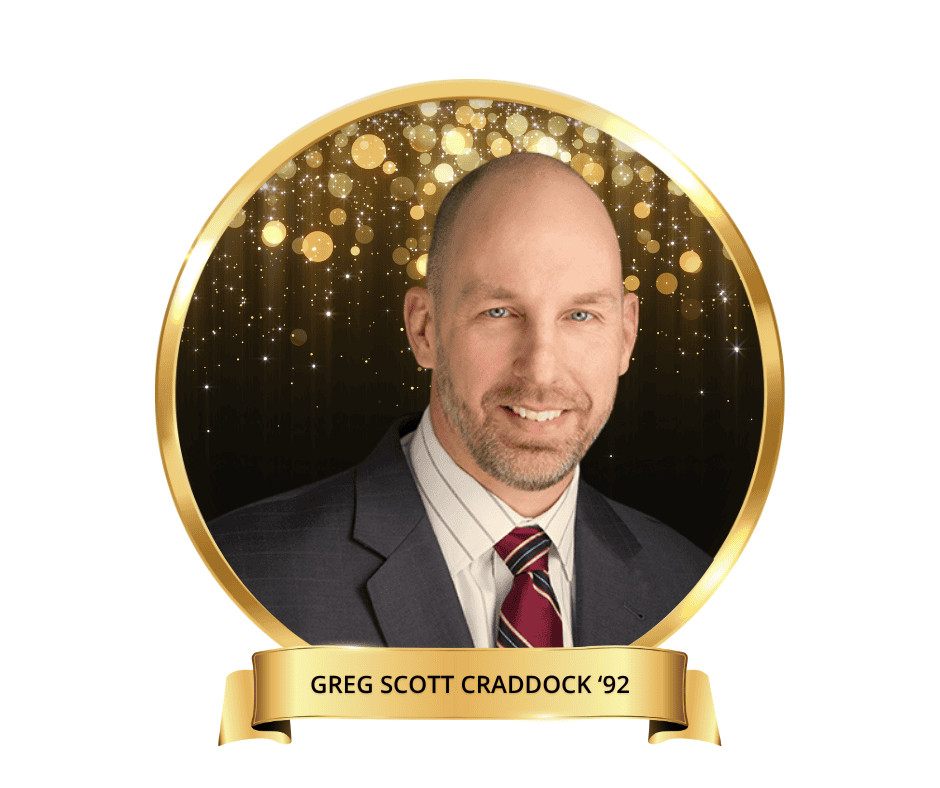 A headshot of Greg Scott Craddock. Wearing a grey suit with a red tie with a black and gold drop.