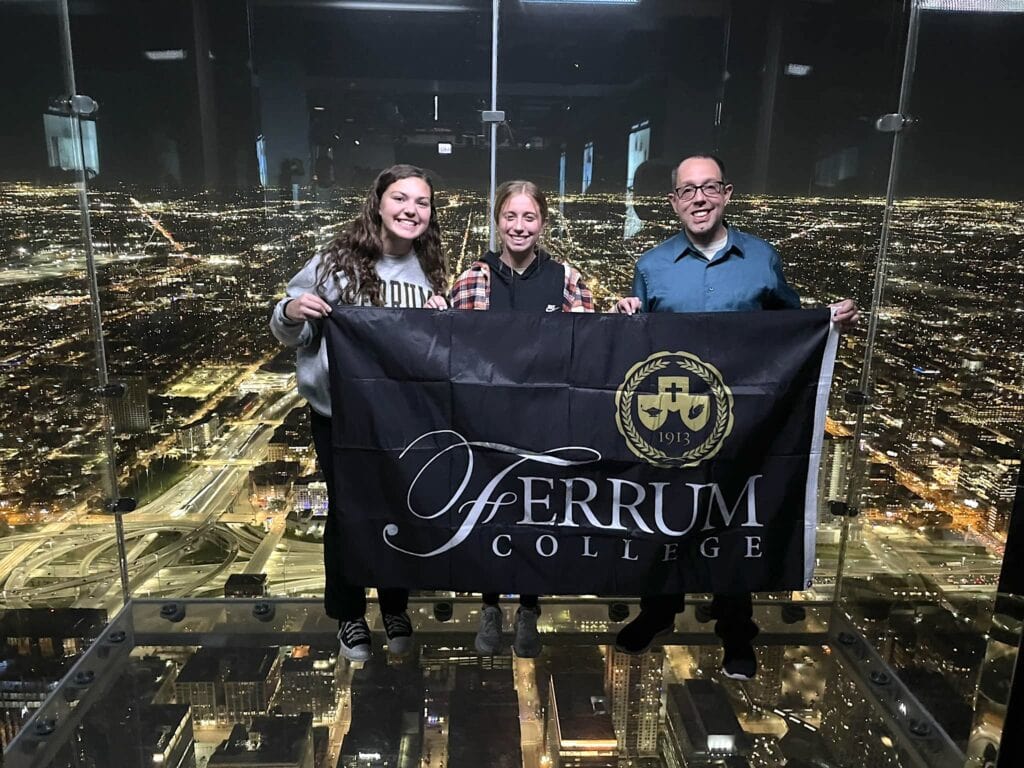 Ed Hally and 2 honors students holding Ferrum College banner in a glass big city overlook with dark night cityscape behind them