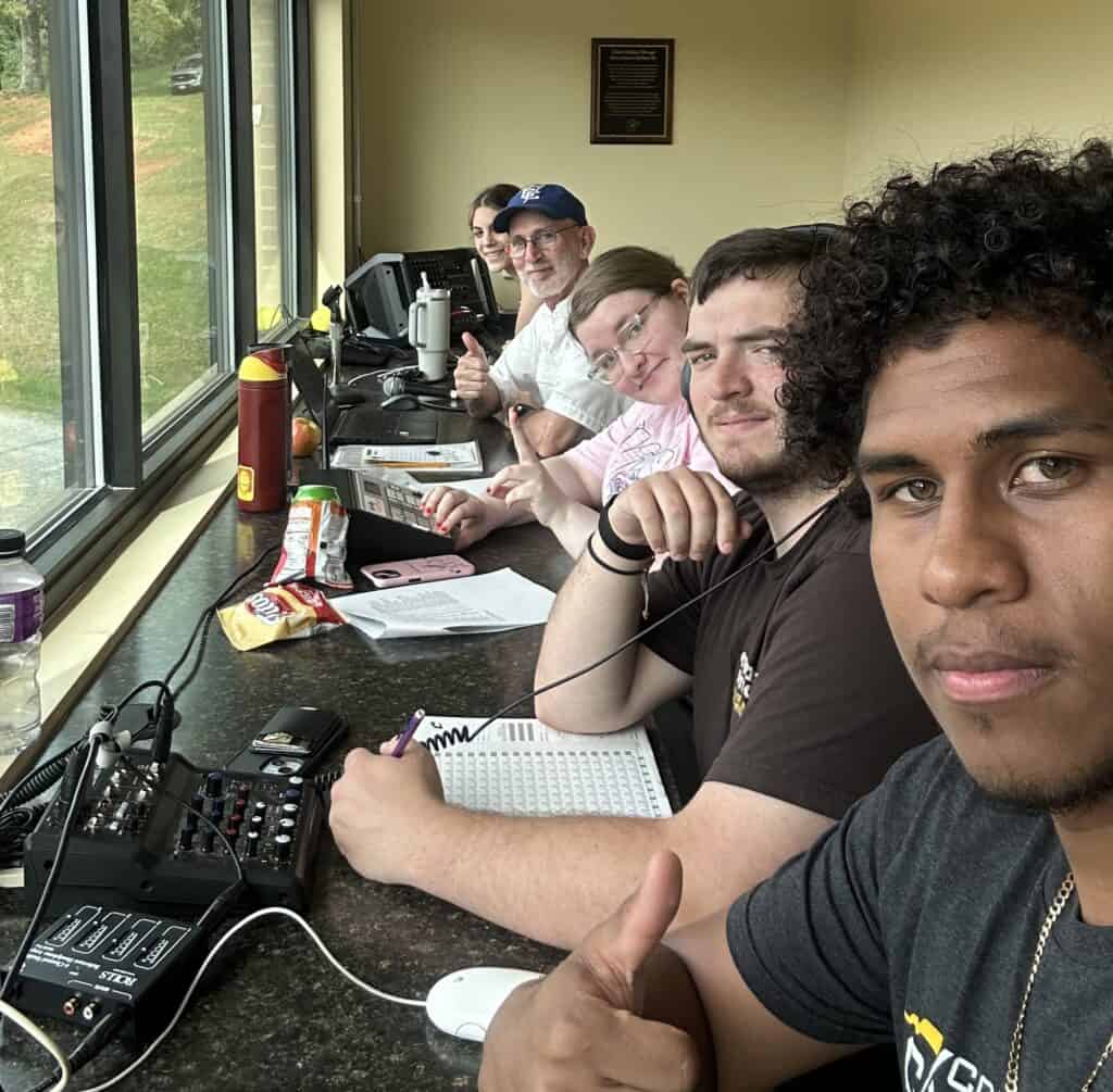 Gary Holden in blue ball cap with beard, with 4 student workers in a row from the side, at the window of an announcers' booth, with a bit of outside showing through windows