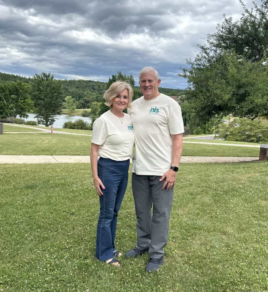Bonnie Huffman and Darrell Huffman in light yellow camp shirts and dark pants on lawn at Ferrum College with sidewalk and Adams Lake in background