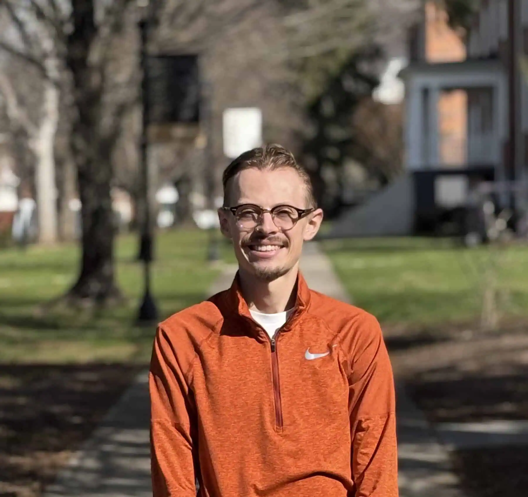 A man with glasses in a orange pullover. Standing in the sunshine.