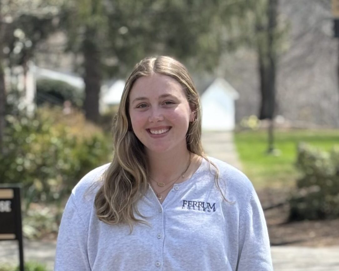 A blonde haired woman standing in the shade wearing a white sweater with Ferrum College on it.