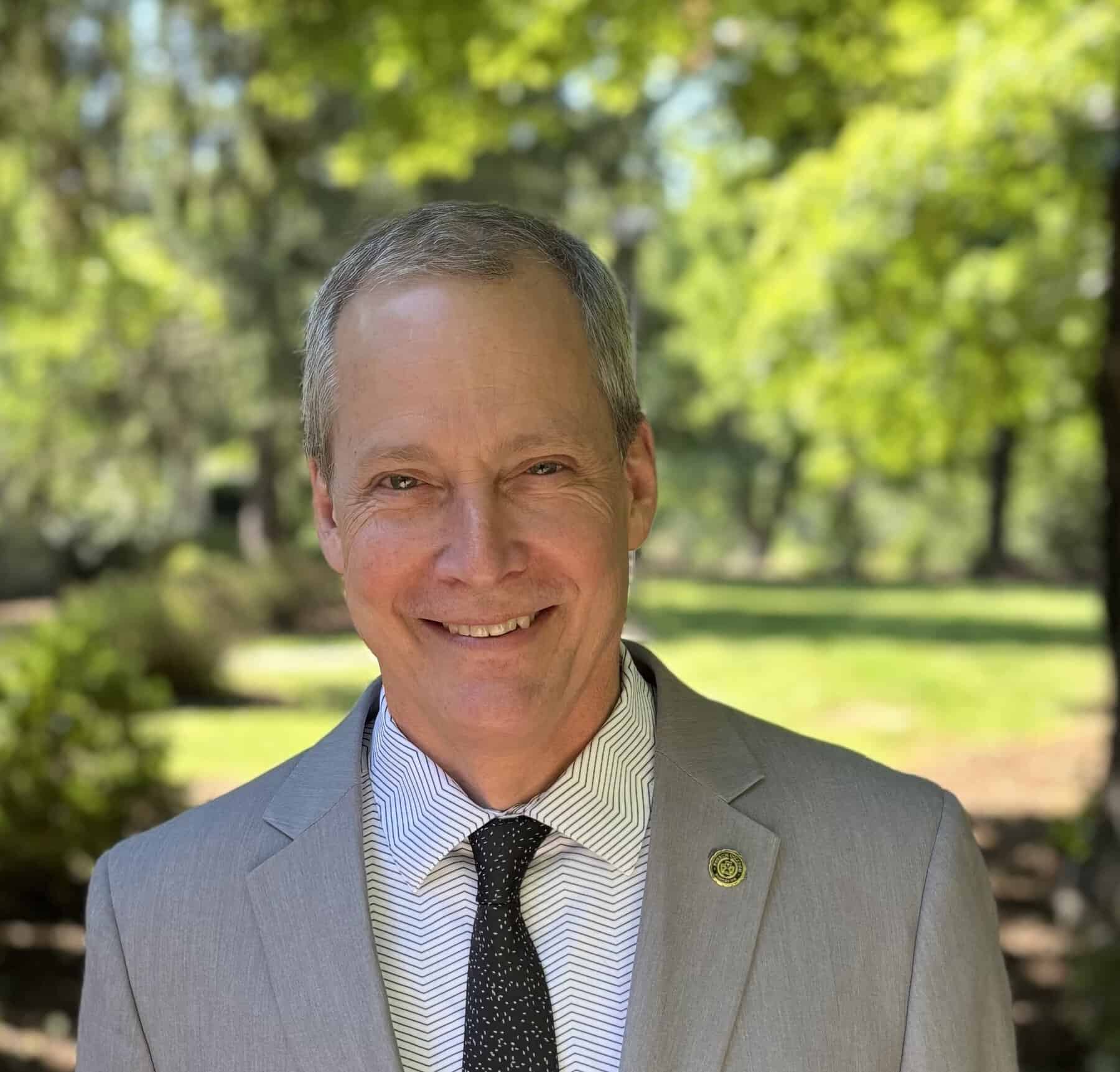 Dr. Richard Grant standing in the shade on a sunny day. Wearing a grey suit, a white shirt with stripes, and a black tie. Smiling at the camera.