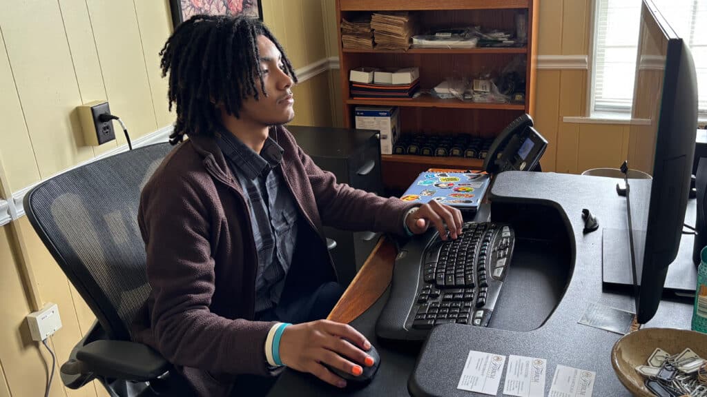A young man sitting at desk typing on a keyboard. He's wearing a brown jacket with a black button down underneath. He has a laptop next to him and a Shelf filled with different things.