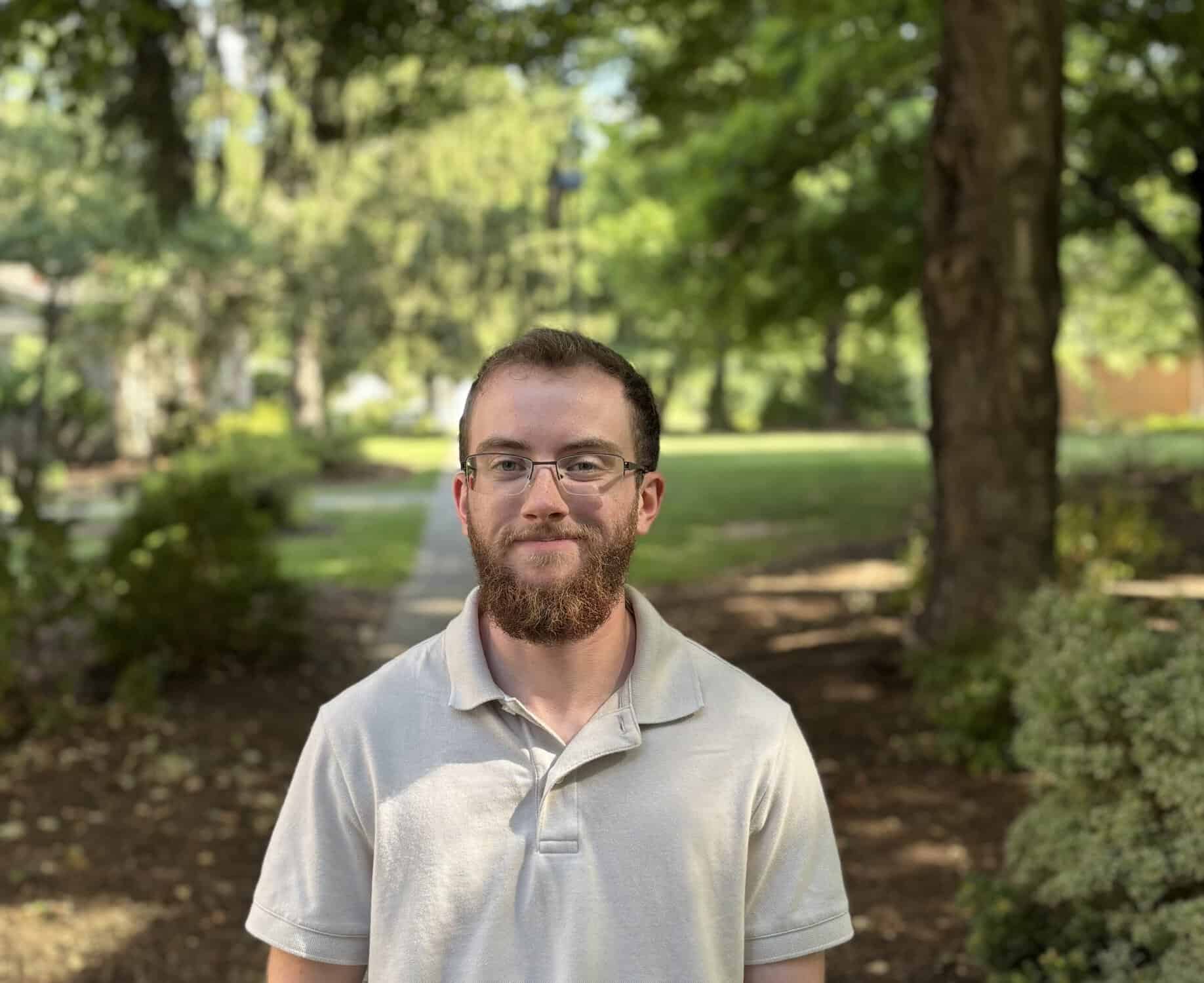 A young man with a beard wearing a gray shirt. He's standing in the shade wearing glasses.
