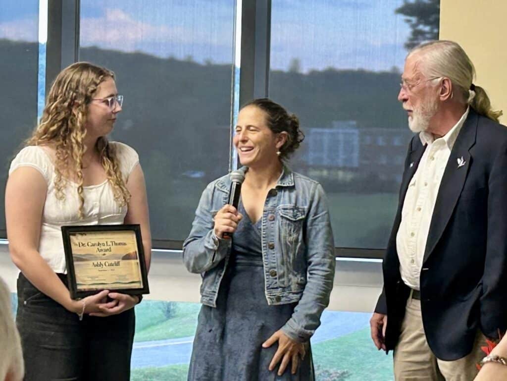 Tall man in dark jacket and white hair standing with short woman in denim jacket and skirt next to college student with long light curly hair, shite shirt, and dark pants, holding a framed certificate in front of large window