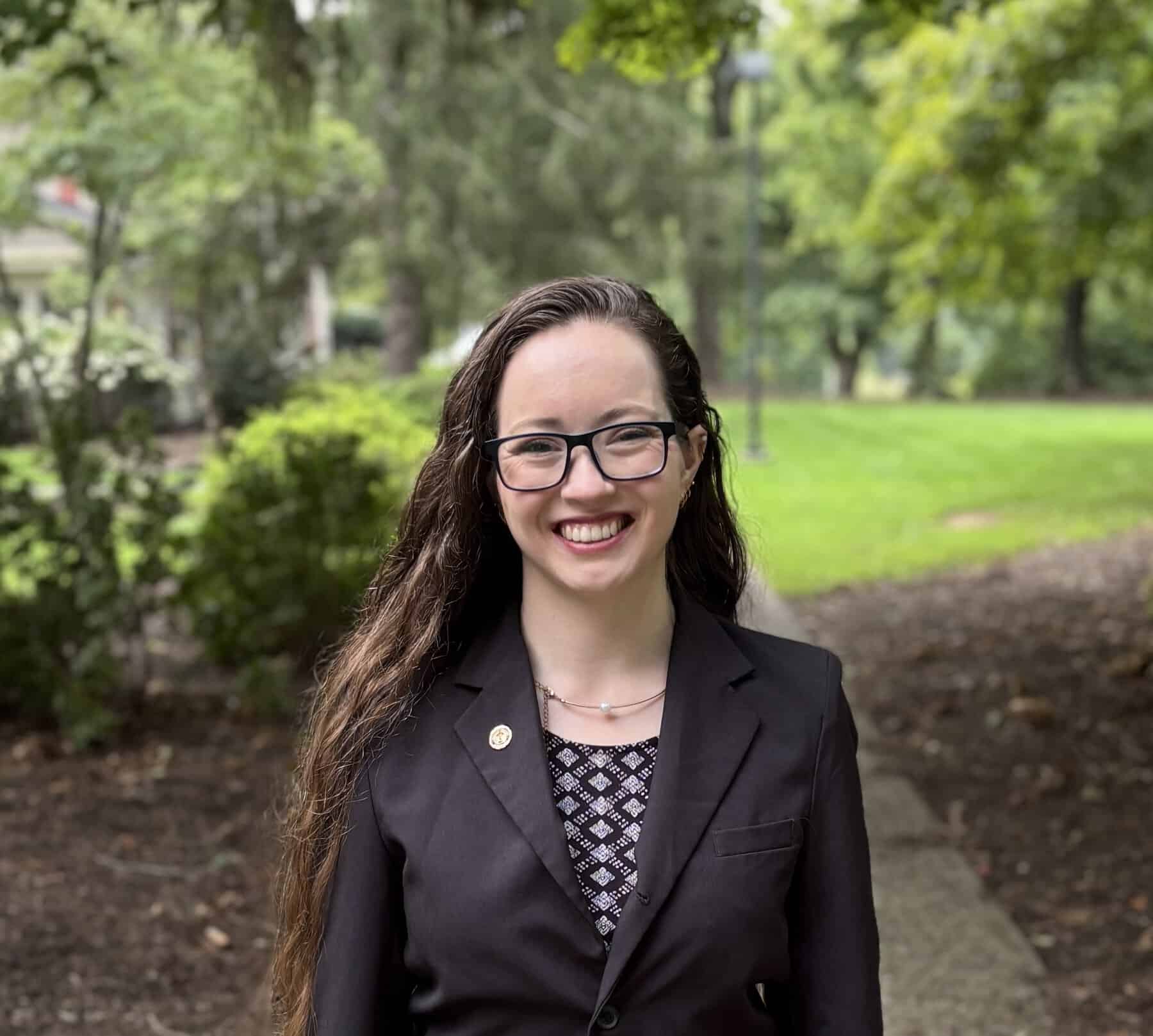 A smiling woman with glasses and long dark hair wears a black blazer with trees and bushes in the background, projecting a warm, confident, professional tone.