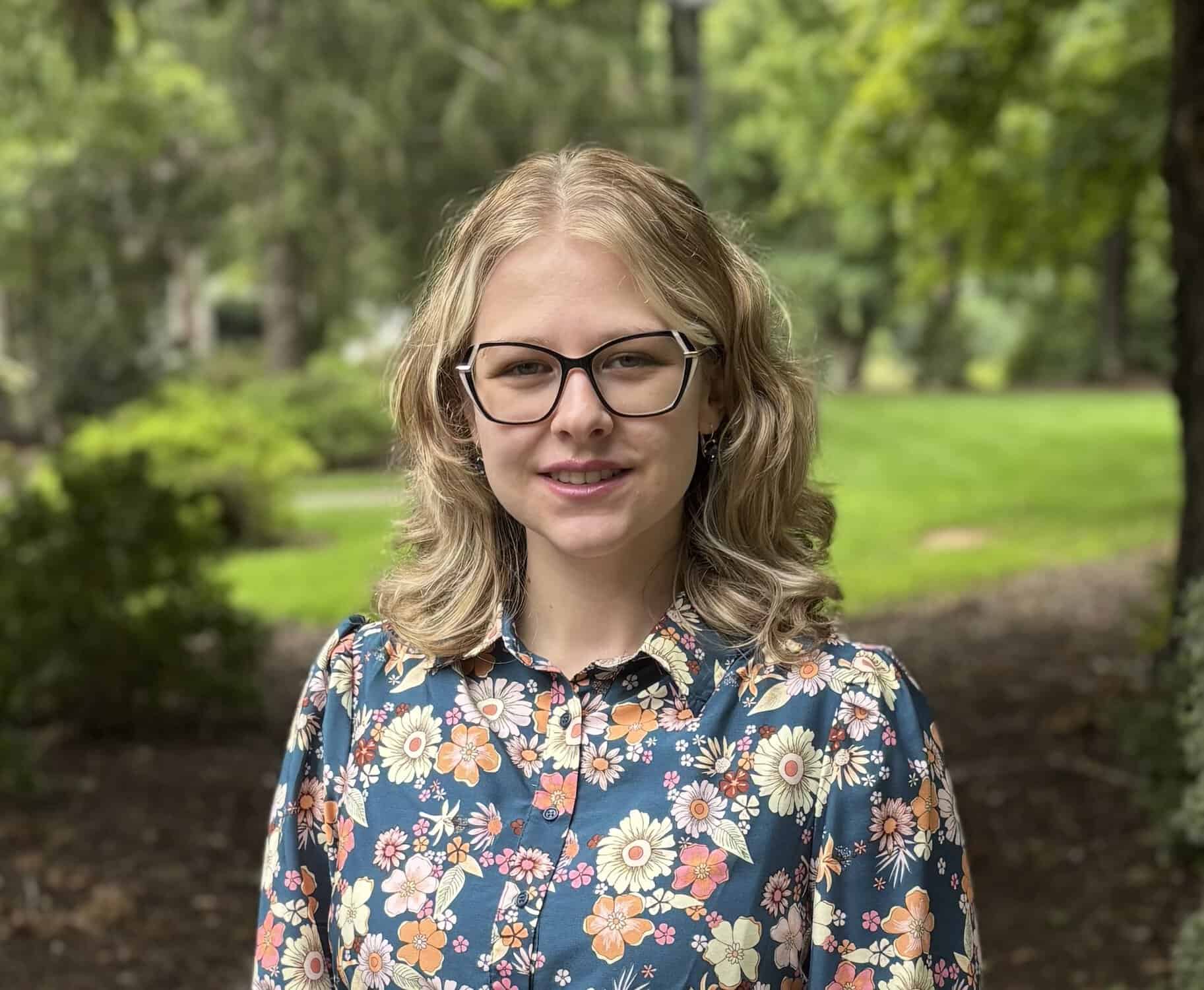 Smiling woman with glasses and wavy blonde hair poses in a colorful floral blouse outdoors, with a lush green park softly blurred in the background.