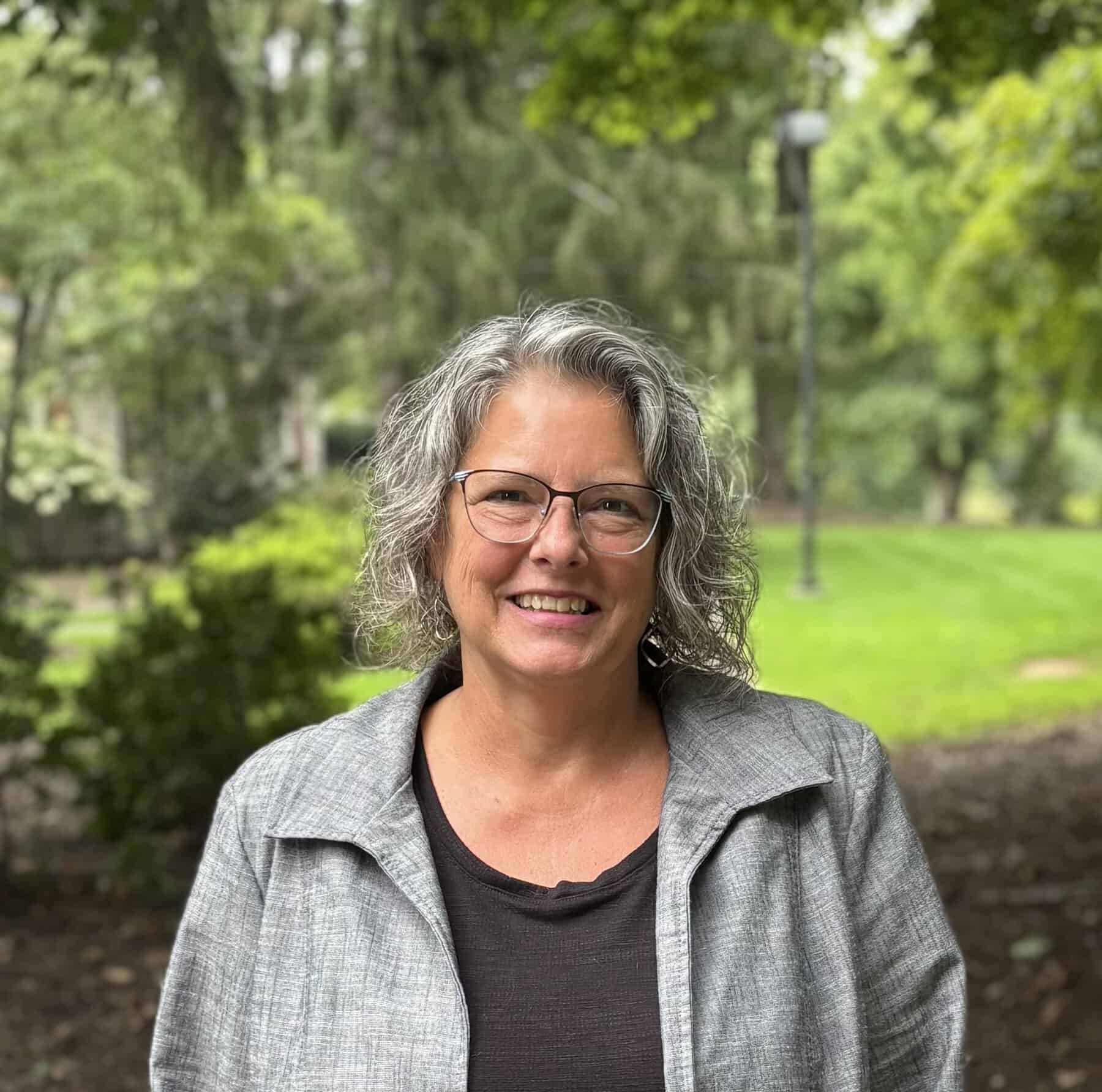 A smiling middle-aged woman with gray hair and glasses wearing a gray jacket with Ferrum College greenery behind her