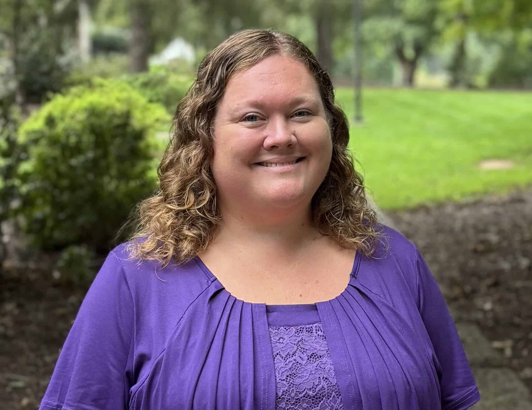 A smiling woman with curly brown hair stands outdoors in a purple blouse, before green shrubs, trees, and a sunny lawn.