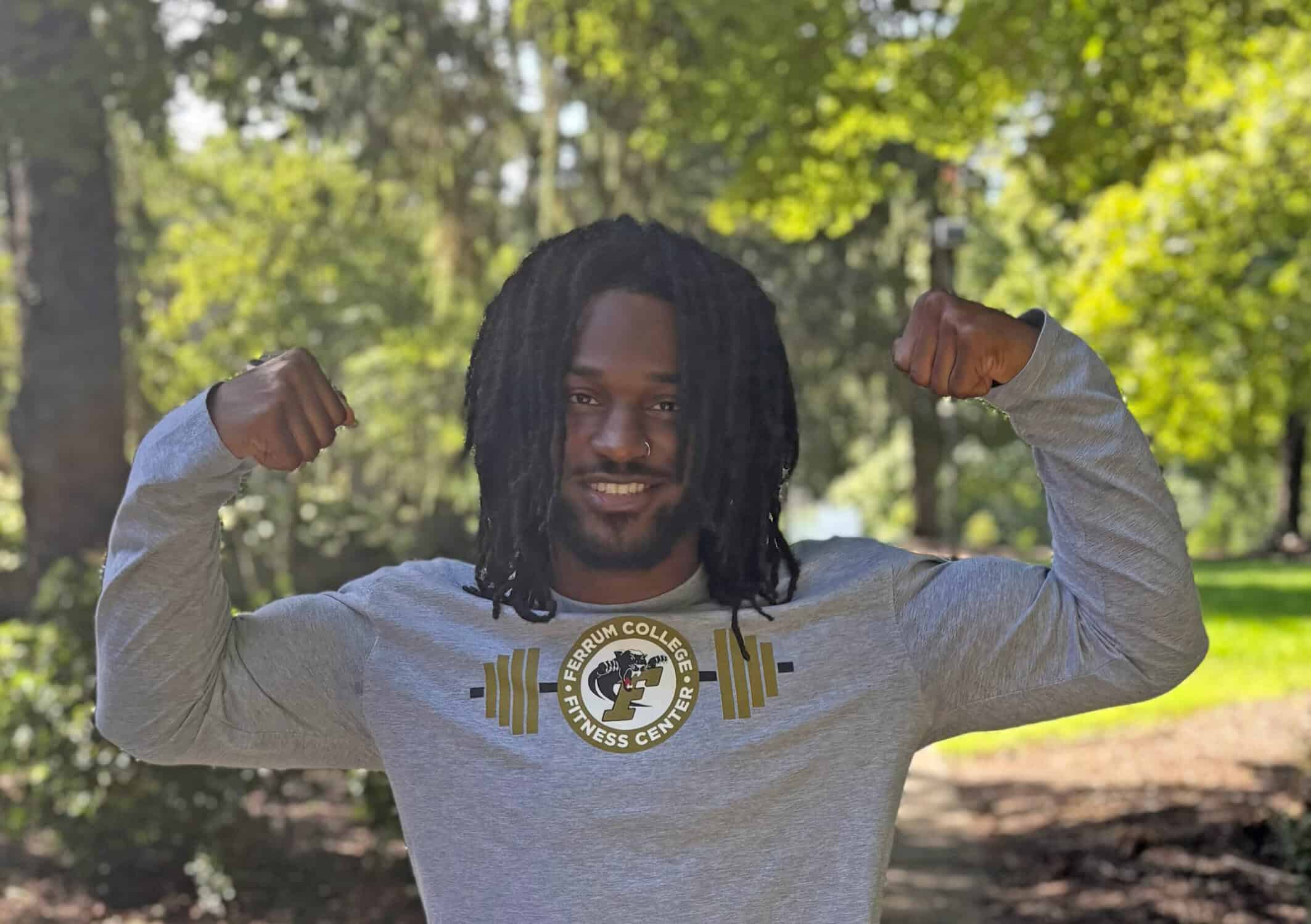 A smiling man with dreadlocks flexes both arms in a park, wearing a gray Ferrum College fitness center shirt; confident, upbeat mood.