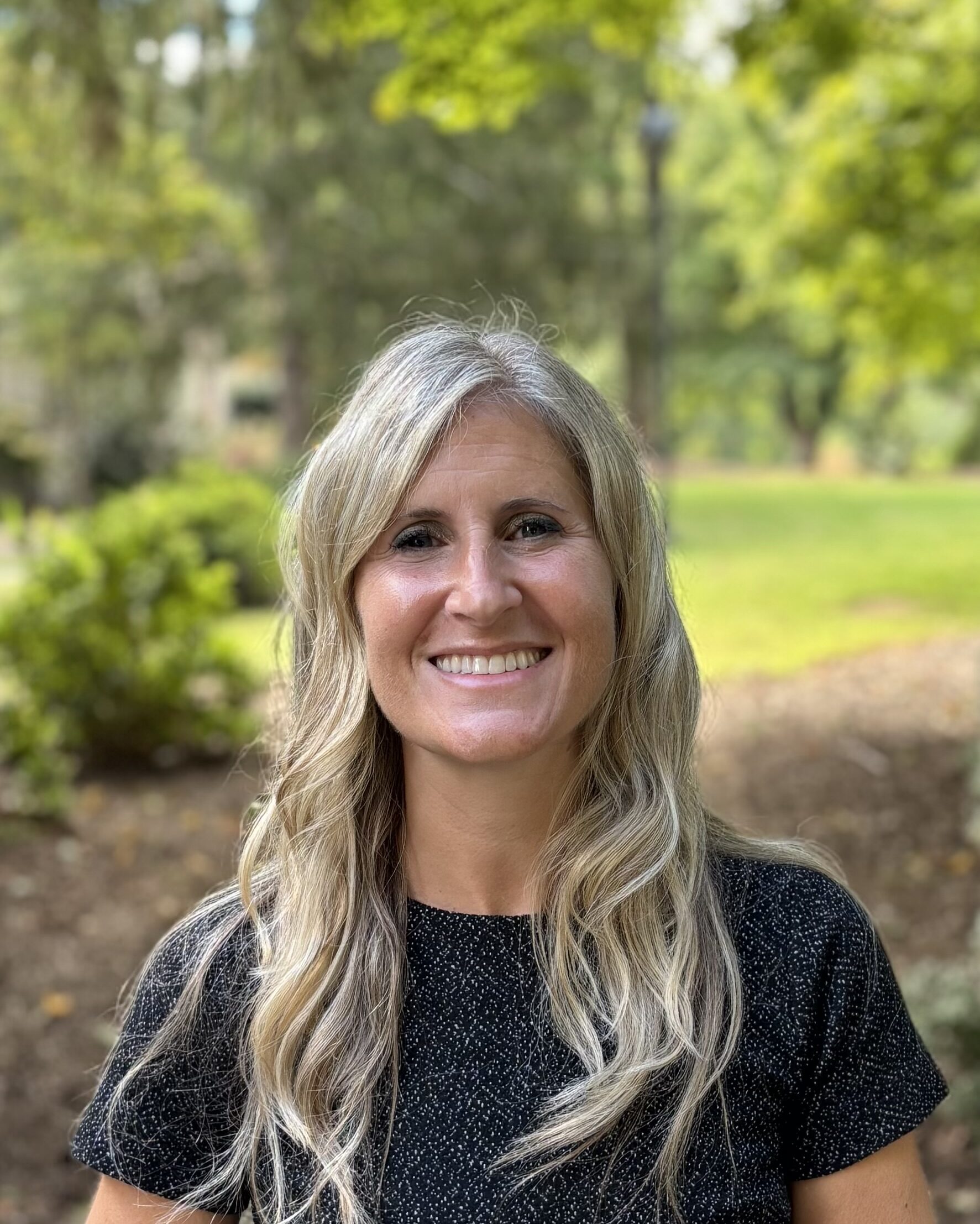 A woman with blonde hair and a black shirt, smiling on a sunny day. The trees and bushes still have green leaves on them.