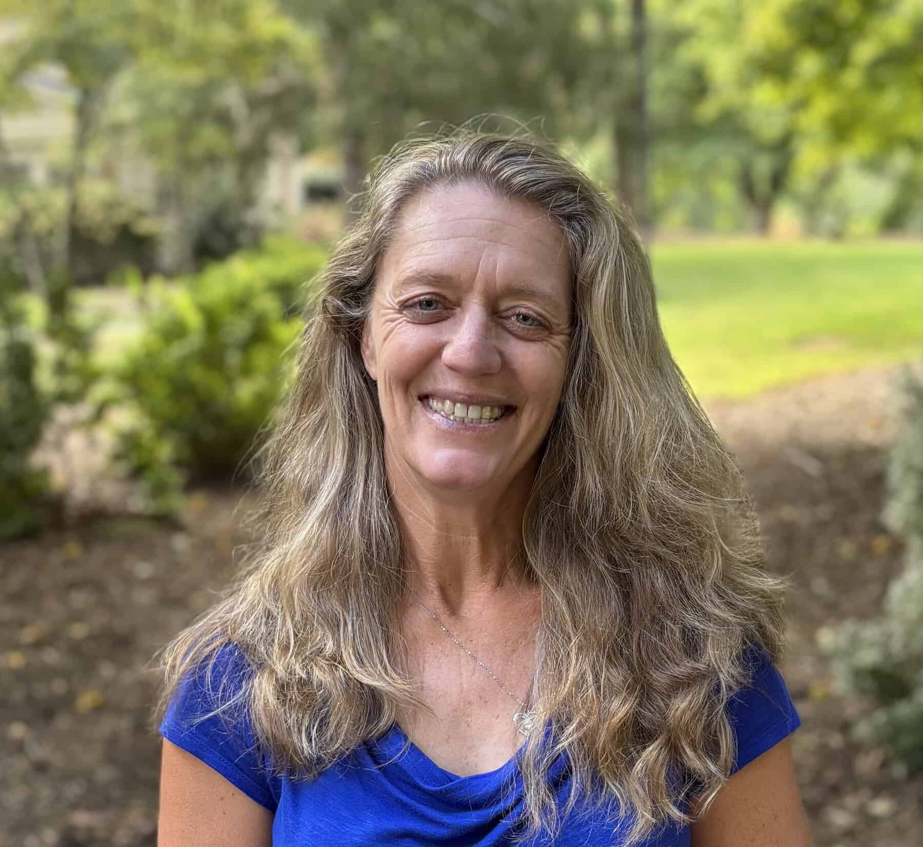 A smiling woman with long, wavy gray-blonde hair and a blue shirt poses outdoors, with leafy trees and a sunlit garden softly blurred behind her.