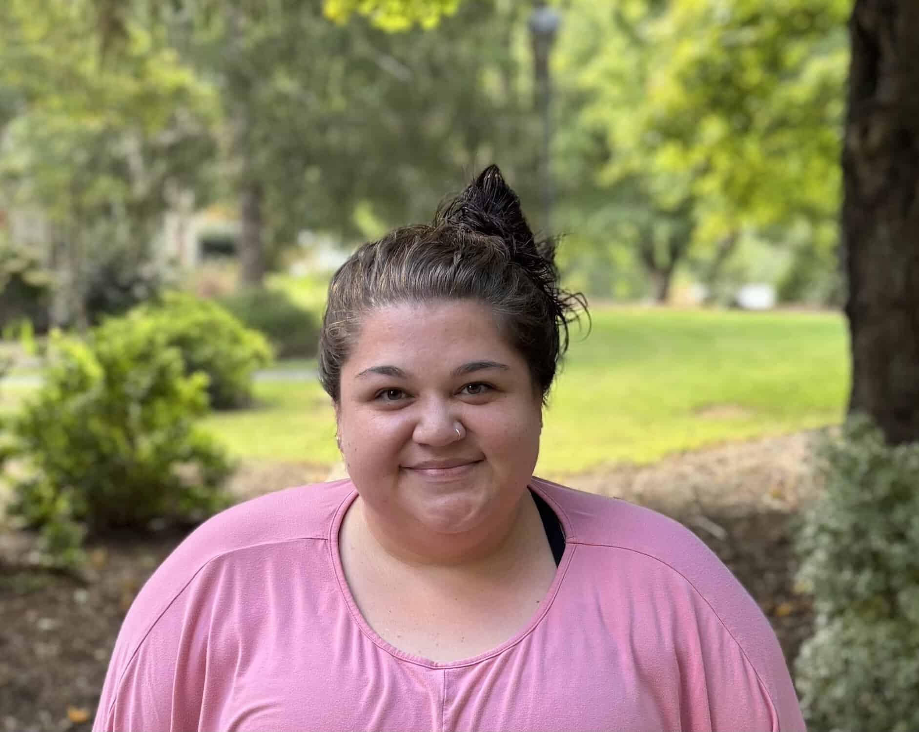 A woman with brunette hair tied up in a bun. She's wearing a pink shirt smiling on a sunny day. Behind her is trees and bushes.