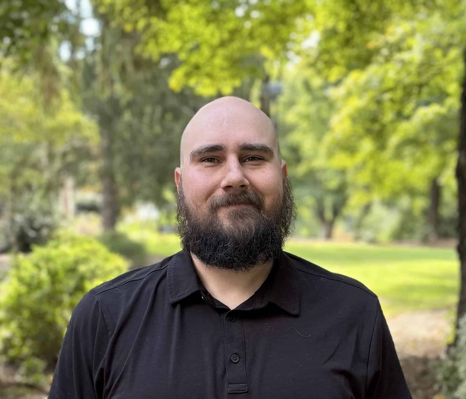 A bald and bearded man smiling. He's wearing a black buttoned up shirt. On a sunny day with the trees and bushes showing green leaves in the background.