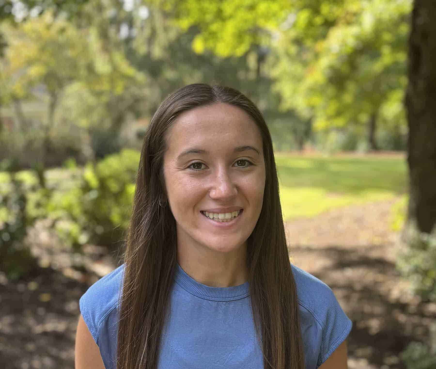 A young woman with brunette hair smiling. She is wearing a blue t-shirt. Standing on the sidewalk on a sunny day with the trees and bushes still having green leaves.