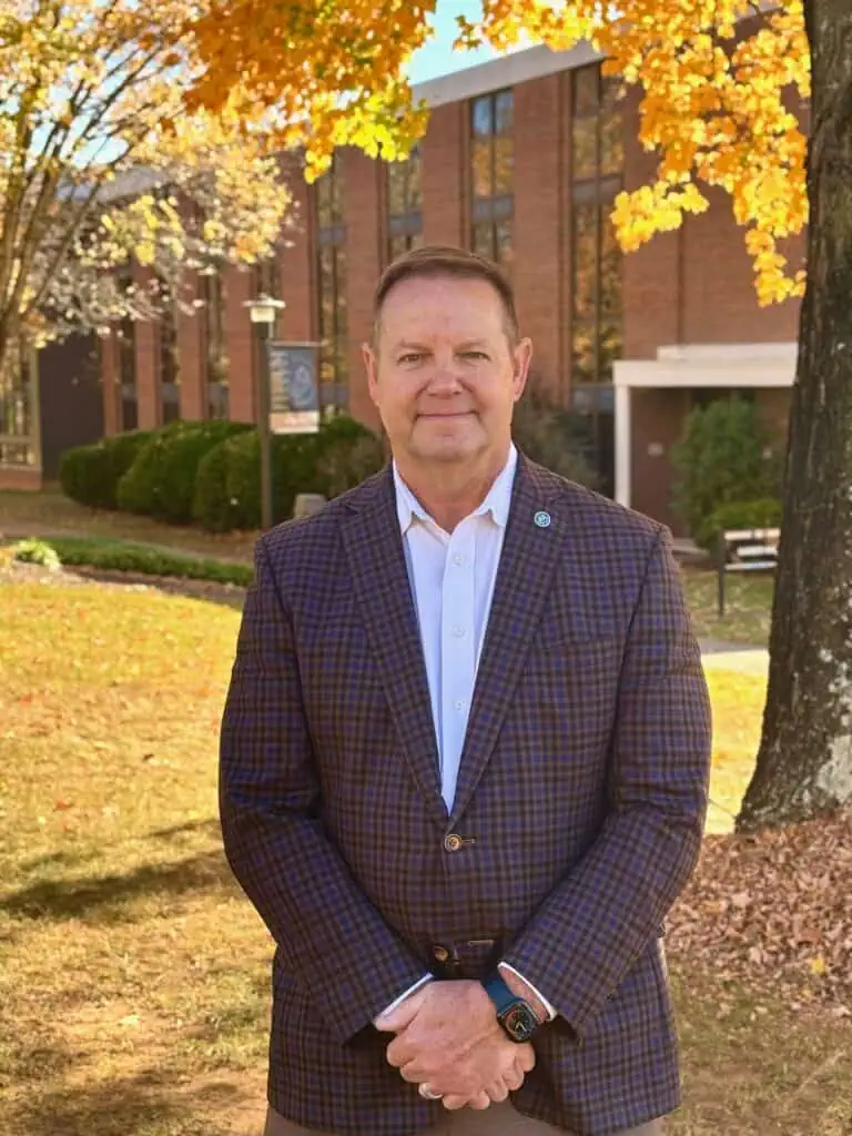 An image of Jim in a suite standing beneath autumn leaves on Ferrum College Campus smiling.
