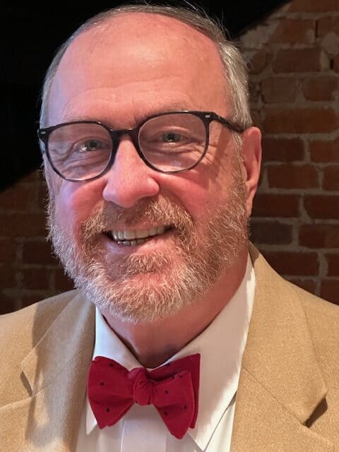 Head shot for Carthan Currin, in tan jacket, white shirt, red bowtie, glasses, with dark background