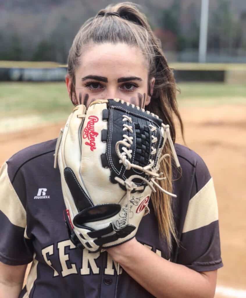 Ferrum College softball player posing on field with mitt up over her mouth, with long brown hair pulled back