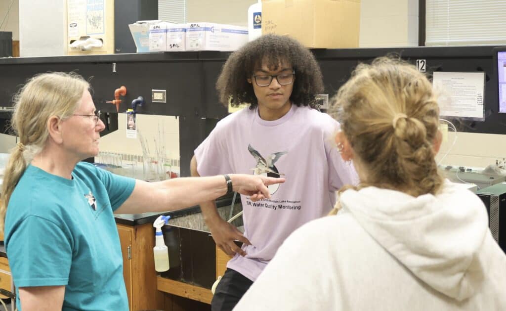 Carol Love in blonde hair and turquoise shirt pointing at something in science lab with two students facing each other on right