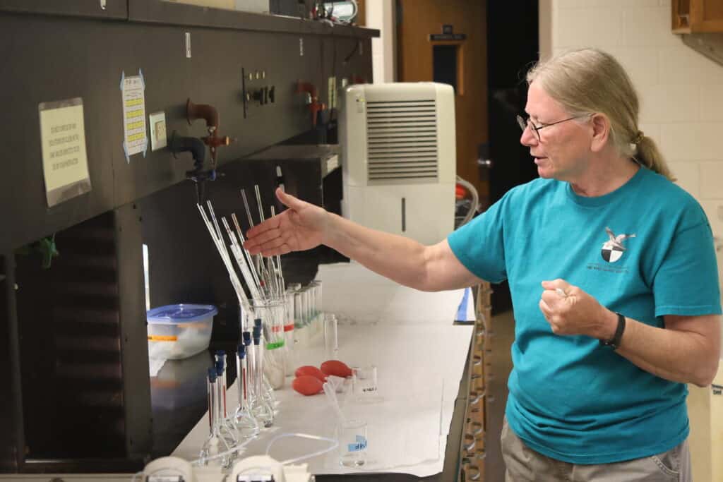 Carol Love in blonde hair and turquoise shirt pointing to something with right arm in science lab full of equipment
