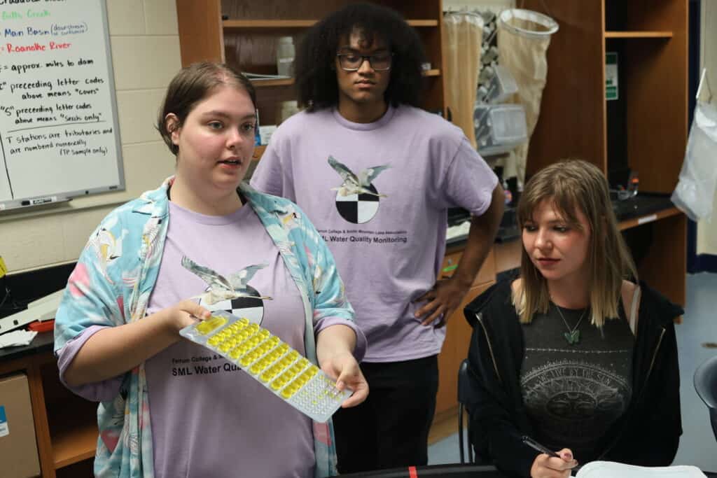 Three students standing and sitting in Garber lab, two with lavender water quality monitoring shirts on. One in front is holding a flat scientific display with many yellow dots on it. Science equipment in back