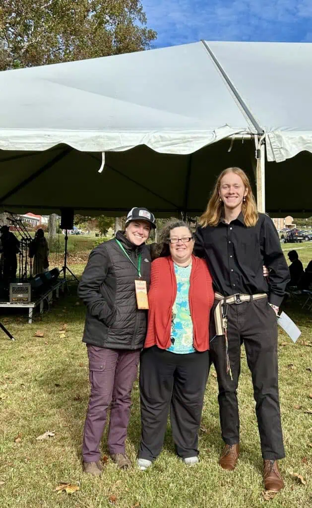 Two members of Orchestra Appalachia in dark colors flanking a shorter woman in a red jacket, dark pants, and multicolored shirt, in front of a white tent at the folklife festival