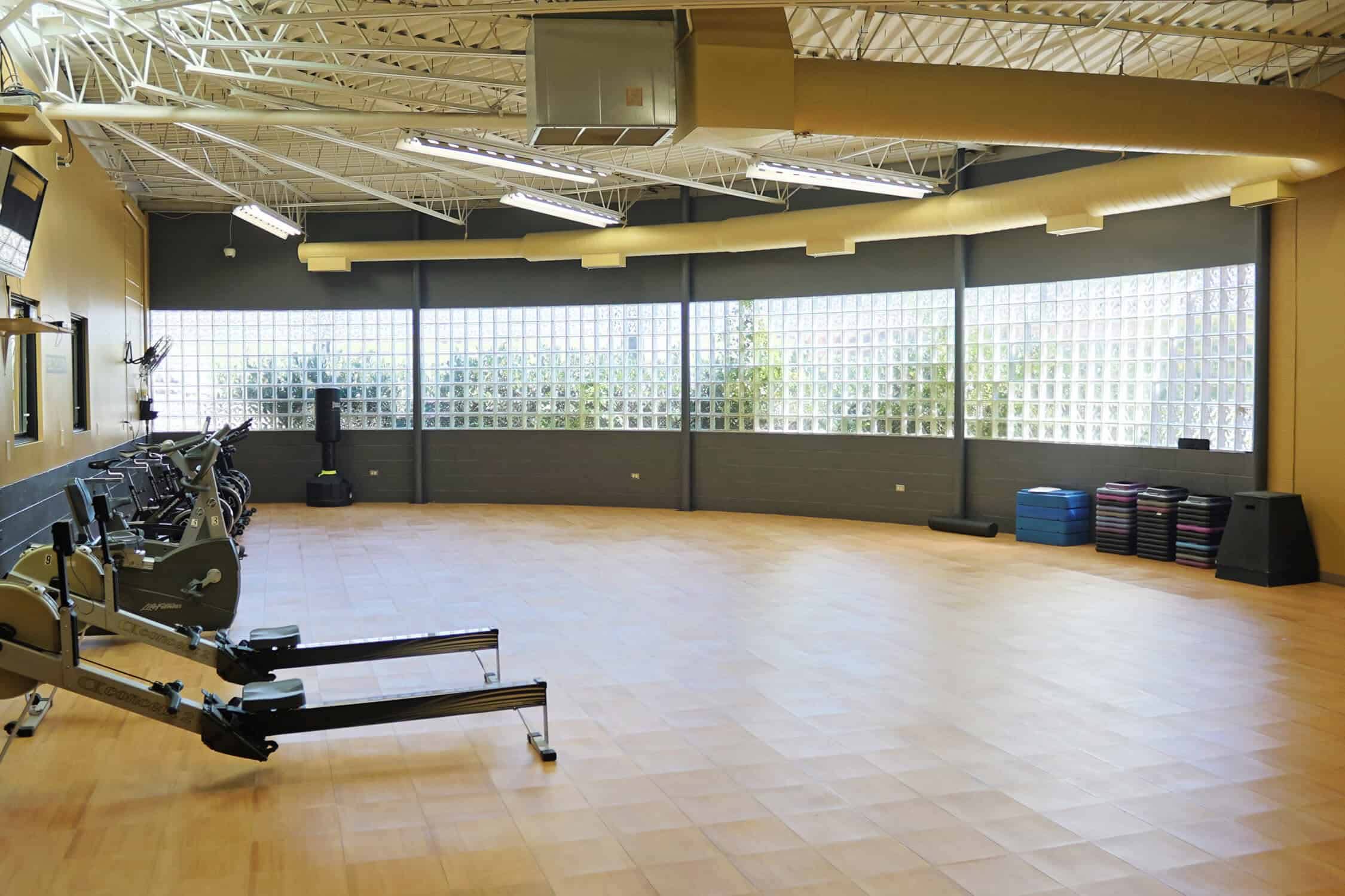The studio with hardwood flooring. There are stationary bikes lined up on the wall to the left there are two row machines next to the bikes. There is a standing punching bag up against the back wall.