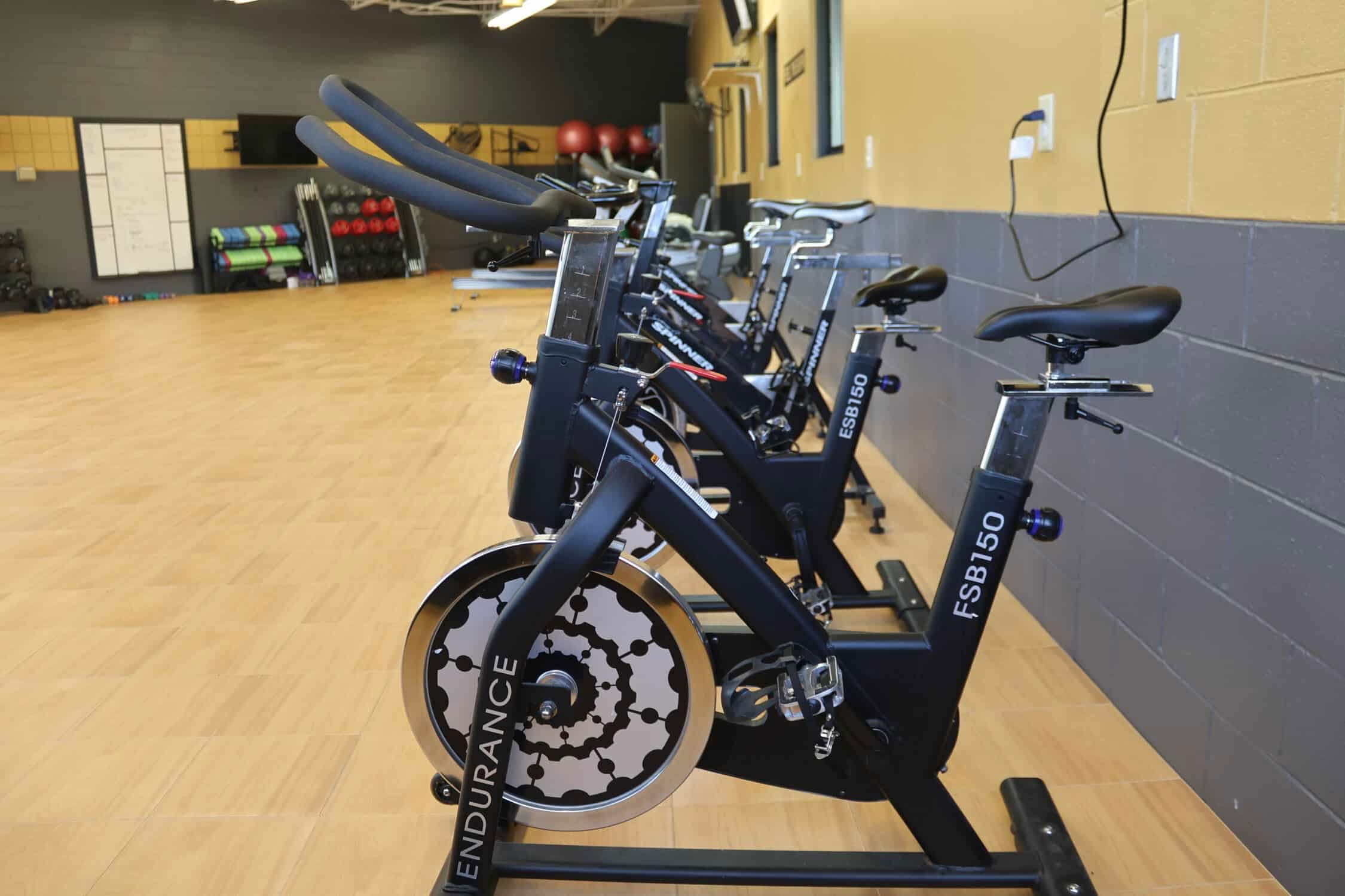 A row of stationary bikes. All lined up next to each other. The bikes have adjustable seats and handlebars. The bikes are sitting on a wooden floor.