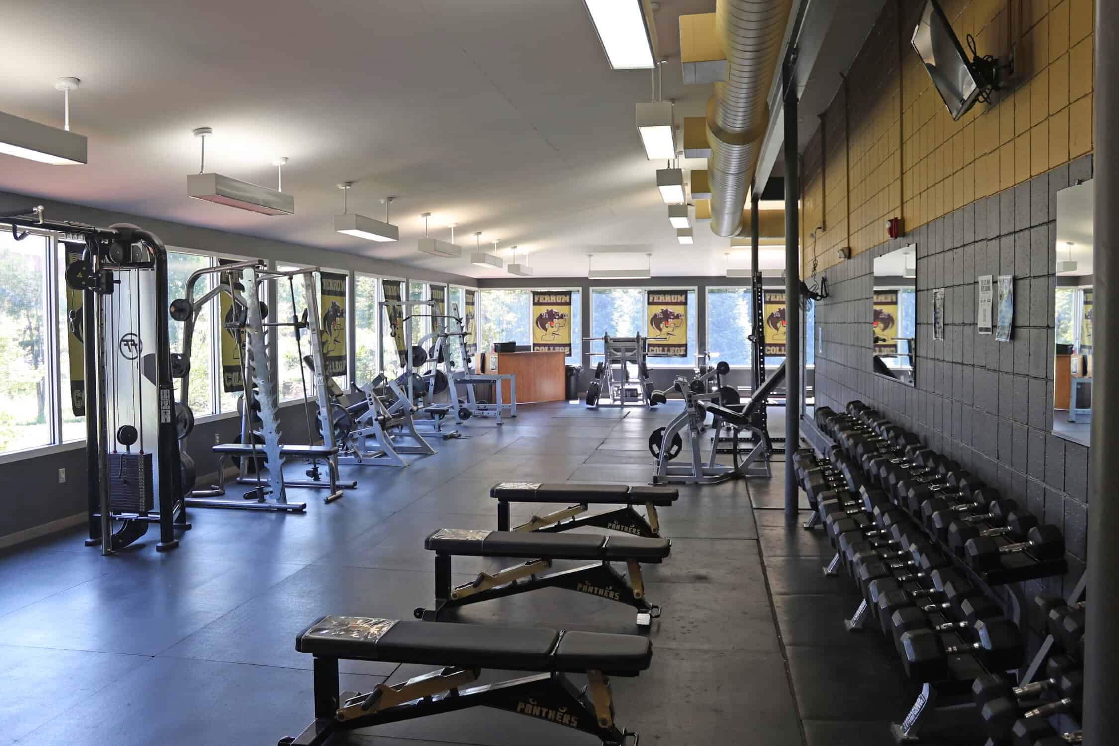 Dumbbell rack up against the right wall. They are organized from lightest to heaviest. Three free weight benches are in front of the dumbbells. A pulldown and smith machine both are on the left wall. Down at the end is a desk with a leg press machine and squat rack next to it.