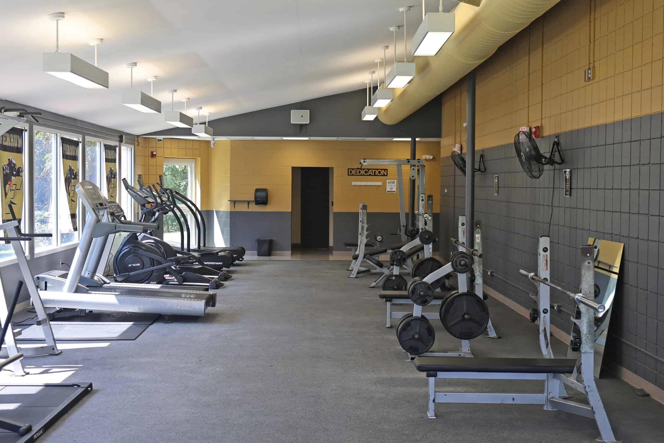 A gym with bench racks to the right and treadmills to the right. The equipment sits on a carpet floor. Weight racks are in between the benches.