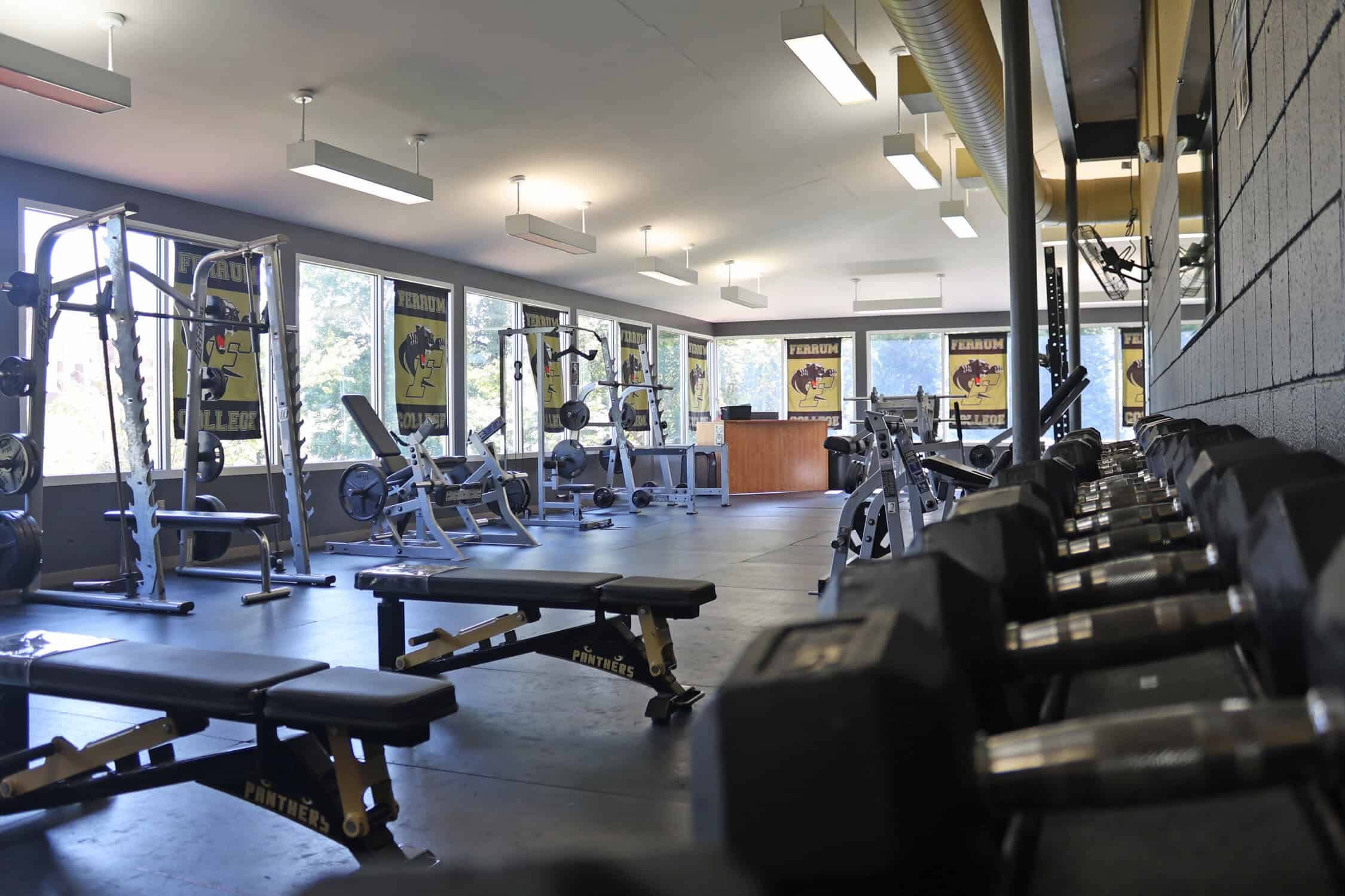 Dumbbell rack up against the right wall. They are organized from lightest to heaviest. Three free weight benches are in front of the dumbbells. A pulldown and smith machine both are on the left wall. Down at the end is a desk with a leg press machine and squat rack next to it.