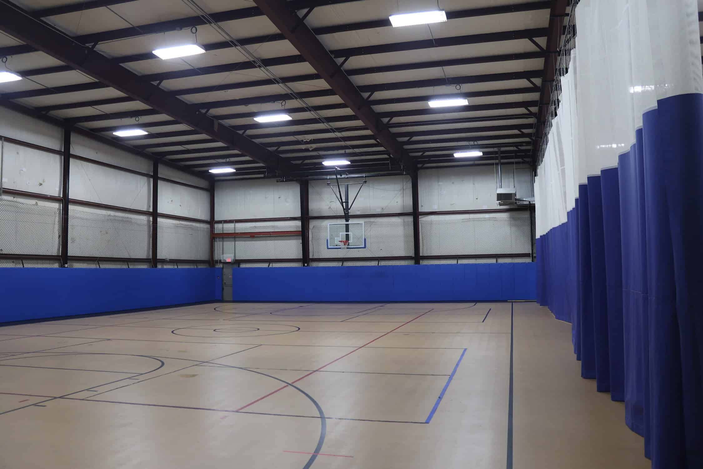 An empty gym with a curtain dividing the other gym floor. There is a basketball hoop at the end of the gym.