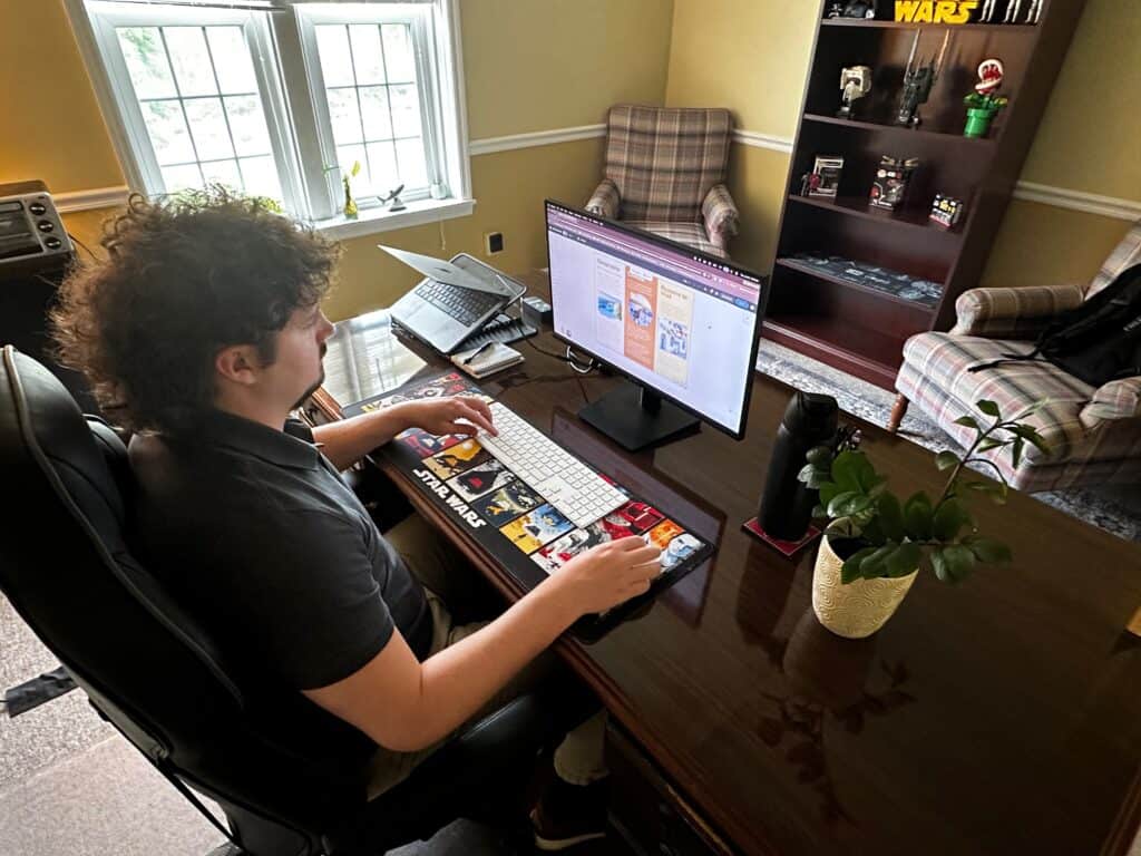 Young man with thick curly hair at a desk working on laptop, with bookshelves and plaid chair and bright windows in the room.