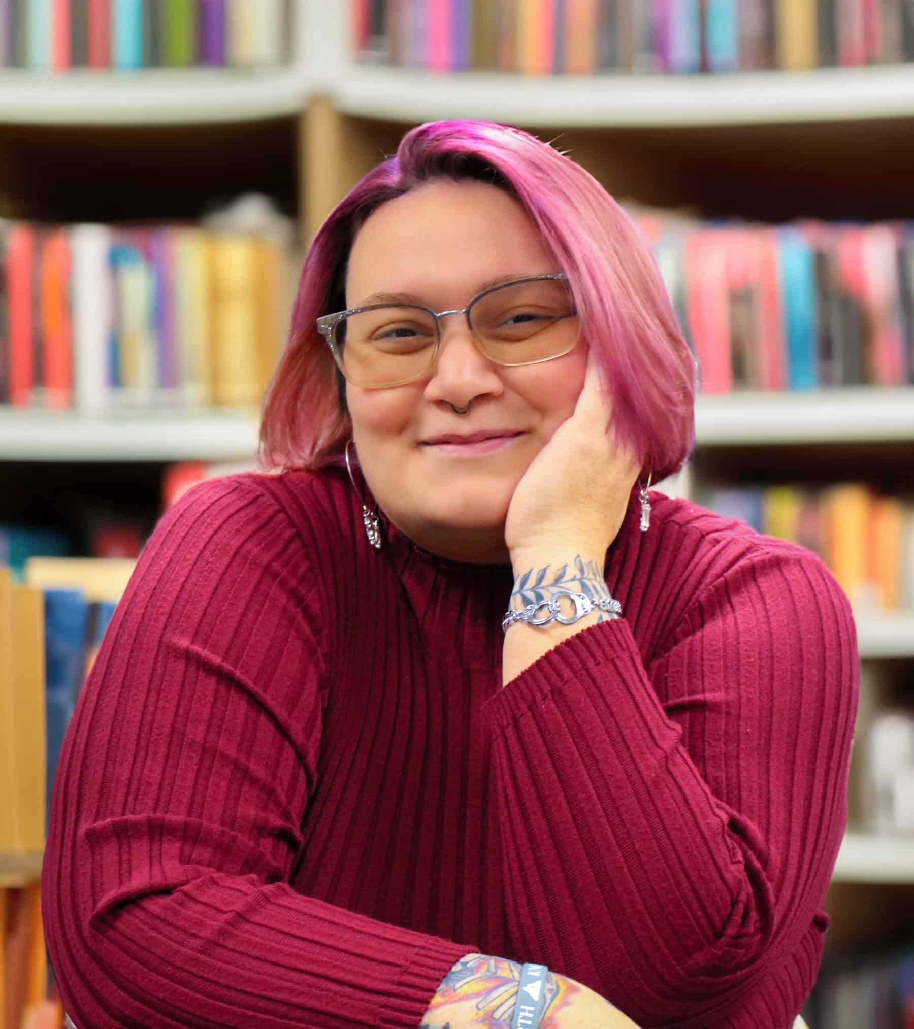 Headshot of Rachel Walton '15 with dark pink sweater and pink hair, wearing glasses in front of shelves of books