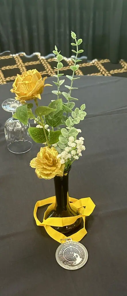 Banquet centerpiece on a black tablecloth with two yellow roses, small white flowers and green leaves, with gold ribbon wrapped around the bottom of a thin vase, and gold Boone Honors Program medallion on the ribbon.