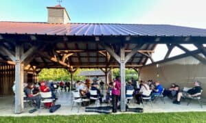 Monday Music Jam in wooden pavilion with roof and blue sky showing, grass in foreground, beige sunshade hanging down on the right, and many people playing musical instruments and sitting around the musicians on white chairs.