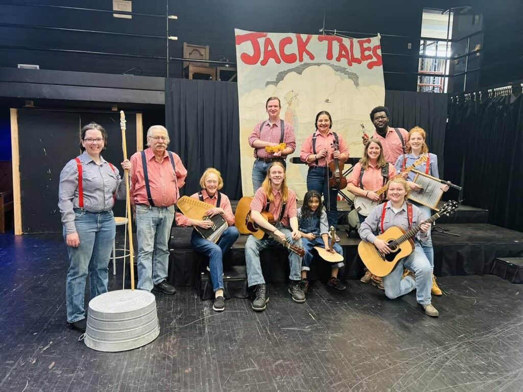 Jack Tale Players ages 8-80 with their instruments in Rex Stephenson Theatre in front of faded Jack Tales backdrop, in red and blue gingham shirts and suspenders and blue jeans, with washtub bass on left, founder Rex Stephenson second from left