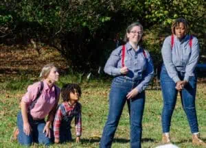 Four Jack Tale Players performing outdoors, two standing in jeans and blue gingham shirts and red suspenders, and two kneeling as animal characters, in red plaid shirts and blue suspenders--one adult and one child