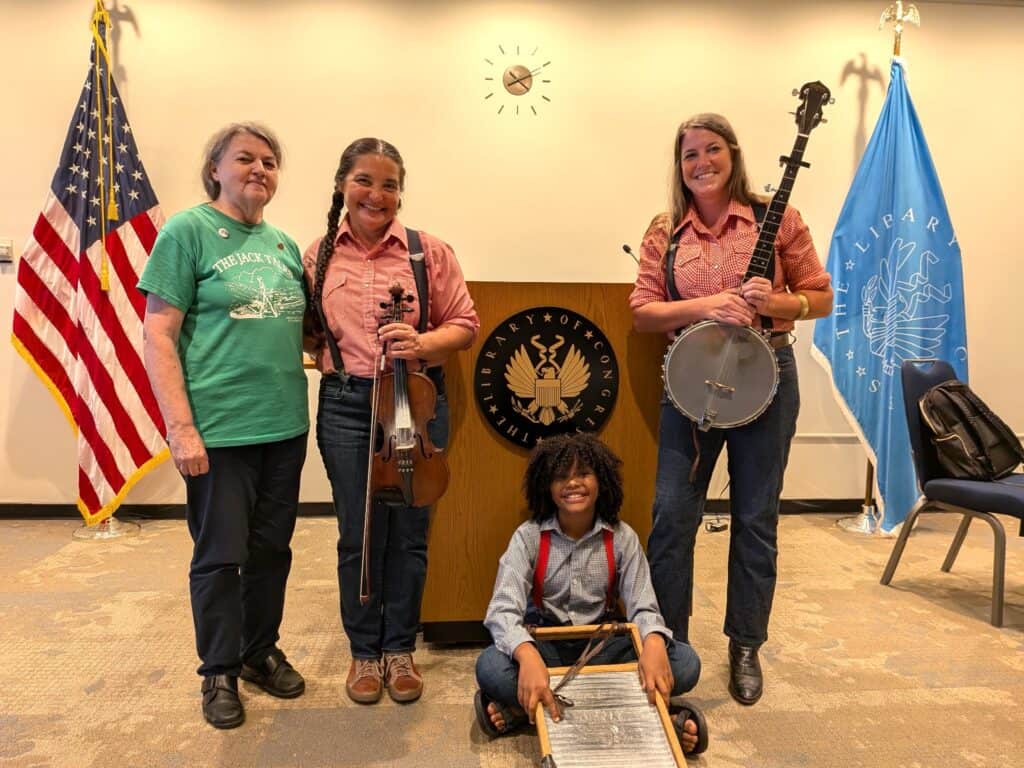Three Jack Tale Players in gingham shirts, suspenders, and blue jeans with traditional instruments, young one sitting on floor and others in front of a Library of Congress podium, with Tina Hanlon on left in a green Jack Tales t-shirt and black pants, with American flag and Library of Congress flag on sides