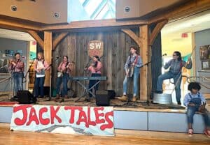 Seven Jack Tale Players in red and blue gingham shirts and blue jeans, playing instruments on a dark stage, with a sign below that says