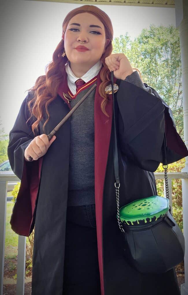 Young woman with long hair standing on a porch in a black Harry Potter school robe, vest, and mostly red tie, holding a wand in front of her chest, and a cauldron like a string bag, that looks like the top is bubbling over with bright green brew.