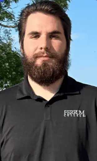 Head shot of Jeremy Quarles in black Ferrum College shirt with dark hair and beard, with a bit of tree and sky in background
