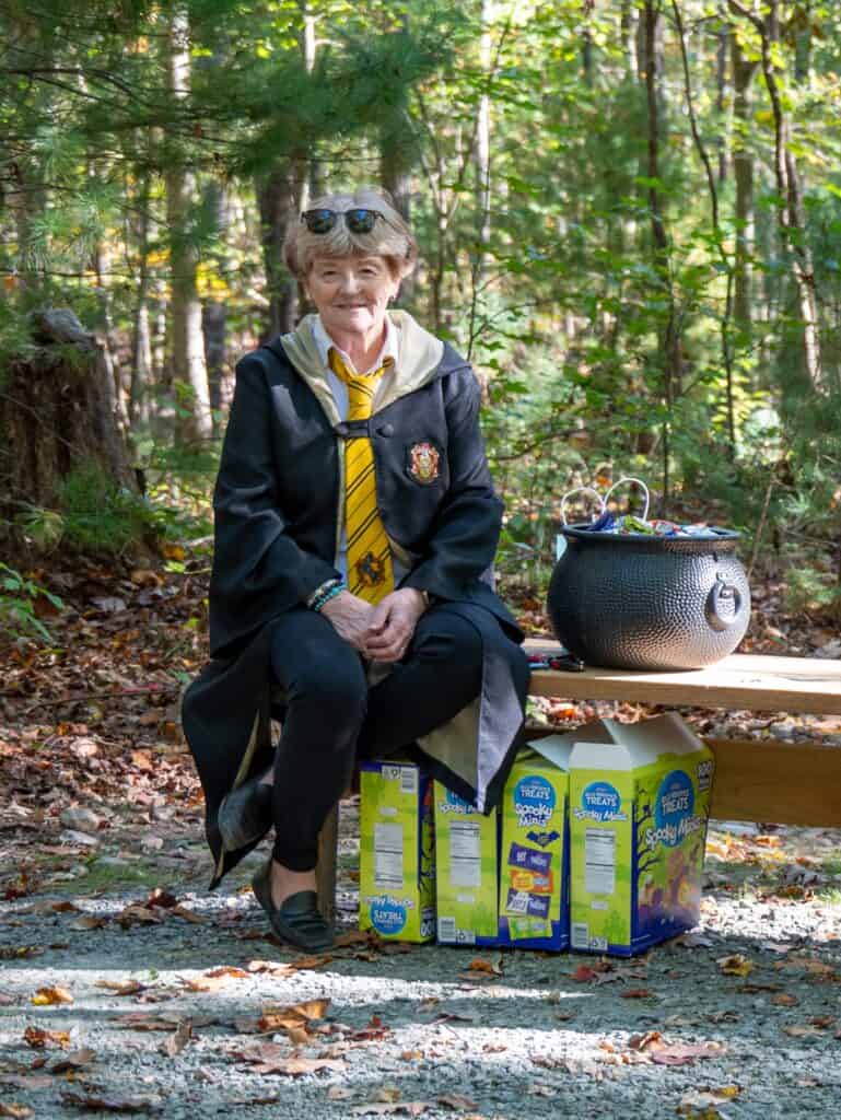 Woman with short red hair sitting on a park bench in woods with a Harry Potter black school robe on and a light colored tie, next to a black cauldron, with boxes full of candy packs at her feet.