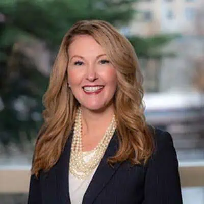 A woman with shoulder-length brown hair wearing a black blazer and pearl necklace smiles warmly. Her expression exudes happiness and positivity, conveying a sense of joy and contentment.